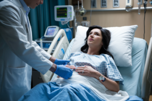 A cinematic shot of a woman in labor, receiving an epidural from a doctor. The woman has dark hair and is wearing a hospital gown. She is lying on a hospital bed. The doctor is wearing a white coat and gloves. He is standing on the left side of the bed. The hospital room has a few medical equipment, including a heart rate monitor.