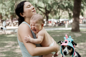 A mother holding her baby in a park, with a humorous touch of a dog wearing a silly hat.