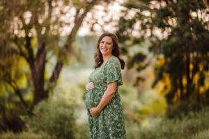 A woman with a post-pregnancy body, smiling and wearing a green dress with a floral pattern, standing outdoors among trees.