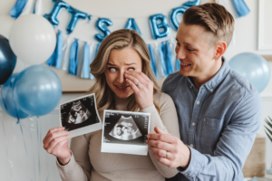 A couple reacting to a pregnancy announcement with balloons and a banner in the background.